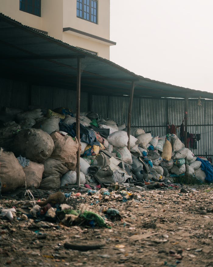 Home Piles of garbage bags stacked in an outdoor waste disposal area near industrial buildings.