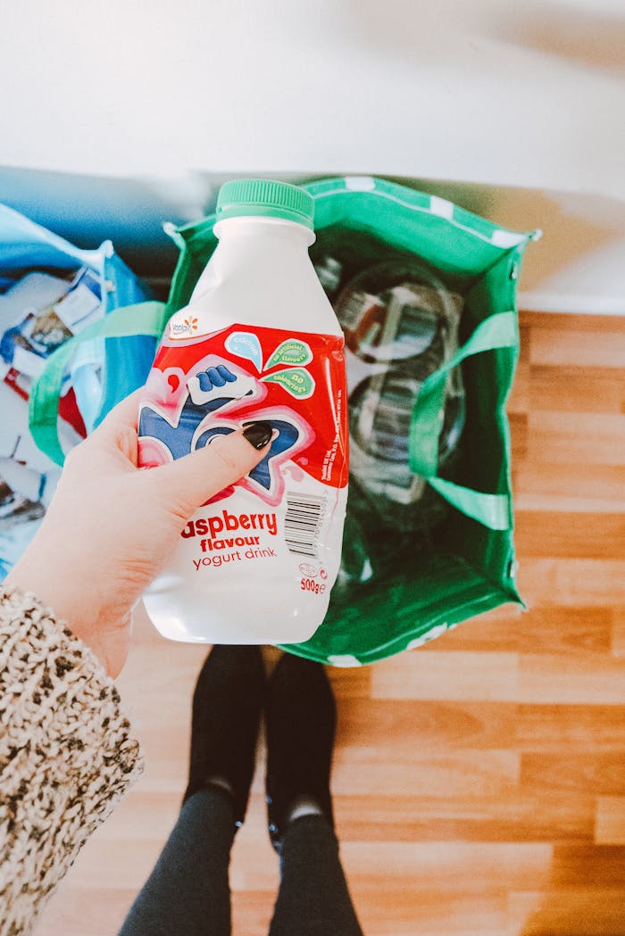 Services A person recycling a raspberry yogurt drink bottle into a green recycling bag.