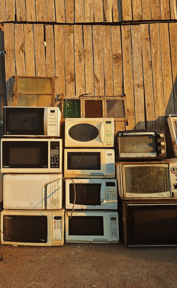 Home A stack of old microwaves placed outdoors against a rustic wooden wall in warm sunlight.